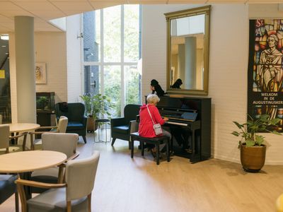Een lichte, grote ruimte met tafels en stoelen en in het midden een vrouw achter een zwarte piano.