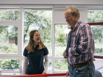 Een man staat tussen de leggers van een loopbrug. Zijn linkerarm zit in een mitella. De fysiotherapeut staat ernaast en lacht.