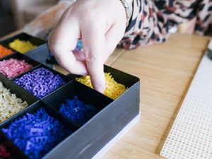 Van dichtbij zie je een hand in een bak met vakjes met gekleurde Ministeck stukjes.