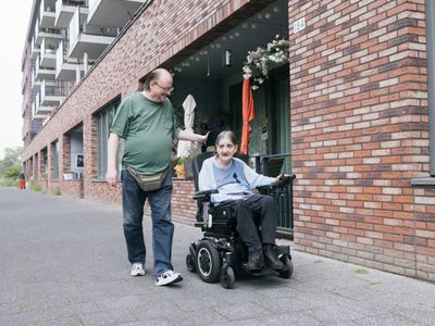 Een vrolijke man met bril wandelt op straat samen met een oudere dame in een scootmobiel.