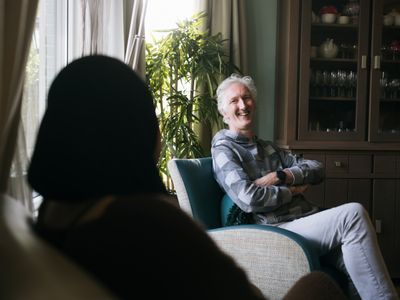 Bewoner zit lachend in stoel met zijn armen over elkaar en is in gesprek met iemand in de gezamenlijke woonruimte