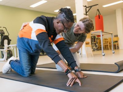 1 persoon is met behulp van een ander oefeningen aan het doen op een mat in de fysiotherapieruimte