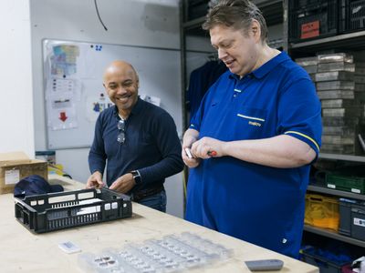 Twee mannen staan in een magazijn bij een houten tafel. Lachend zijn ze bezig met prijskaartjes van producten.