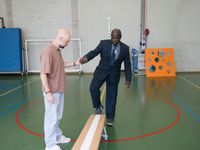 Een man in sportkleding ondersteunt een man in pak in een sportzaal. De man in pak loopt over een gym bank.