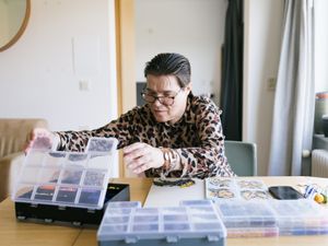 Een persoon met panterprint blouse en een bril zit aan een tafel in een kamer. Op tafel liggen dozen met vakjes en een bord met Ministeck.