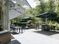 Een terras met tafels, stoelen, bloembakken en parasols tussen veel groen.