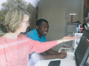 2 personen zitten achter de computer en en de ene persoon wijst iets aan op het scherm