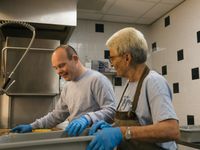 Een man en vrouw doen de afwas in de grote keuken. Ze dragen handschoenen.