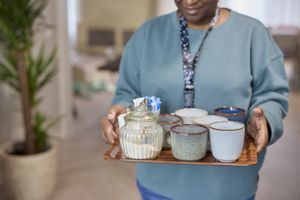 Persoon in een blauwgroene trui die een houten dienblad vasthoudt met zes keramische mokken en een glazen suikerpot
