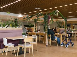 Een eetzaal met houten tafels en stoelen en over een rij stoelen heen staat een groot houten frame in de vorm van een huis, begroeid met planten.