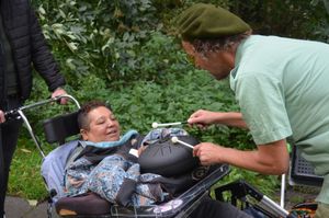 Dame zit buiten in rolstoel en kijkt geïnteresseerd naar een klankkast op haar werkblad dat bespeeld wordt door een muzikant.