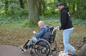 Een jonge man met een pet duwt een oudere dame in een rolstoel vooruit in een groene omgeving. Ze kijken naar de camera.