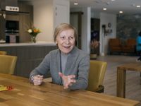 Een kwieke oudere dame met kort haar en lichtgrijs vest zit aan tafel en kijkt met vriendelijke blik naar iemand buiten de foto