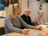 Een vrouw met blond haar in een paardenstaart en bril op haar hoofd zit aan tafel naast een oudere vrouw met bril. Voor hen op tafel ligt een legpuzzel en de vrouwen lachen naar elkaar.
