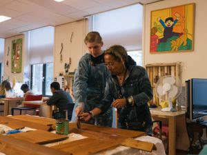 Iemand helpt een ander met het lakken van houten planken. Op de achtergrond doen 2 cliënten een activiteit aan een tafel.