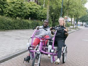 Jonge man met kort zwart haat zit met een grote lach naast een begeleider met blonde paardenstaart p een duo-fiets.