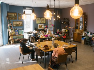 Een gezellig verlichte, gekleurde ruimte met grote vierkante tafels waaraan ouderen zitten en veel kleurrijke kunst in de ruimte.