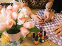 Twee jonge handen houden de hand van een oudere vast en lakken de nagels van deze hand met lichtroze kleur. Op de voorgrond verschillende potjes nagellak