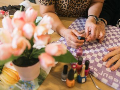 Twee jonge handen houden de hand van een oudere vast en lakken de nagels van deze hand met lichtroze kleur. Op de voorgrond verschillende potjes nagellak