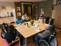 6 mensen waarvan 4 in een rolstoel zitten aan een grote lichte houten tafel met koffie en een krant.