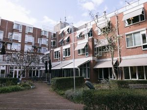 Gebouw van 5 etages van rode baksteen met lichte zonneschermen en tuin met bankje en parasols ervoor