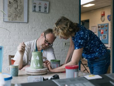 Een man met kort haar en bril kijkt naar een groene keramieken pot, met een kwastje met groene verf in zijn hand. . Een begeleider staat naast zijn tafel en kijkt ook naar de pot.