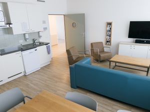 Een kamer met witte muren, een keuken, een zithoek met blauwe bank en een televisie. De deur naar de gang staat open.