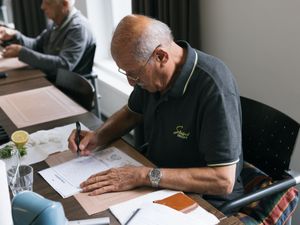Een oudere man zit aan tafel en zit geconcentreerd woorden door te strepen op een woordzoeker.