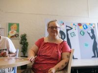 Vrouw met rood shirt, lang blond haar en een bril en tattoos van sterren op haar arm zit in de kantine en kijkt blij de camera in