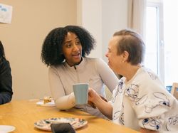 Twee personen zitten aan tafel te lunchen. 1 persoon lacht en heeft een blauwe mok in de hand