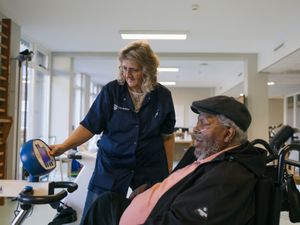 Een man en een vrouw zijn in een sportruimte. De man zit in een rolstoel bij een sportapparaat. De vrouw drukt op een knop op het apparaat.