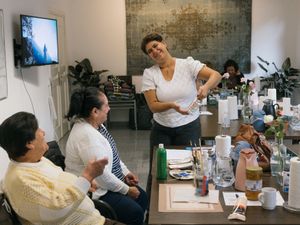 Een vrouw staat bij een tafel met een geschilderd potje in haar hand en kijkt heel trots naar twee oudere dames die aan tafel zitten. Op tafel liggen allerlei schilderspullen.