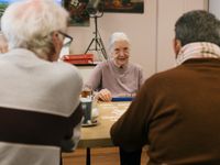 Tussen de ruggen van twee mannen links en rechts zit aan de andere kant van de tafel een oudere vrouw met een kop thee en een bordje met Rummikub stenen.