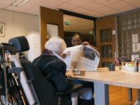 Een dame met kort grijs haar en donkerblauw colbert zit in een rolstoel aan tafel met een kop koffie. Een zorgmedewerker zit ook aan tafel en houdt lachend een krant voor haar op.