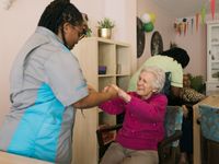 Zorgmedewerker met zwart ingevlochten haar en bril houdt de handen vast van een dame met grijs kort haar en donkerroze trui die vrolijk kijkt en helpt haar uit haar stoel