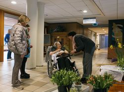 In de galerij met op de grond bossen bloemen geeft een bloemenverkoopster een oudere vrouw in een rolstoel een bosje bloemen en ze lachen naar elkaar. Naast hen staan een zorgmedewerker in blauw werkuniform en een vrouw in een grijze jas.