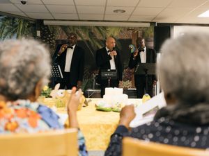 Drie mannen in net pak met strik zingen voor een zaal mensen. We zien ze midden in beeld over de schouders van twee oudere dames.