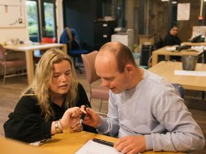 Een man zit aan een tafel met daarnaast een blonde vrouw. Zij helpt hem met tekenen.