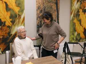Een vrouw staat naast een oudere dame op een stoel en kijkt lachend naar haar. Aan de muur hangen grote posters van herfstbladeren.