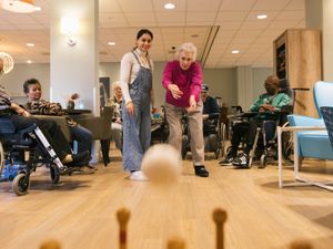 Dame met kort grijs haar en fuchsia trui gooit een witte bal richting kegels. Drie ouderen in rolstoel en een jonge vrouw met lang bruin haar kijken toe