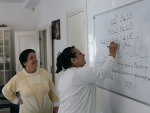 Twee oudere dames met donker haar kijken naar een whiteboard aan de muur en schrijven woorden op in het Syrisch en Nederlands.