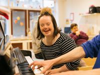 Meisje met zwart-wit gestreept truitje aan zit lachend naast iemand die piano speelt