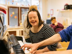 Meisje met zwart-wit gestreept truitje aan zit lachend naast iemand die piano speelt