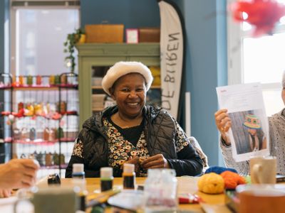 Dame met gebloemde blouse en witte baret zit aan tafel en kijkt lachend in de camera terwijl haar buurvrouw een foto omhoog houdt van een model met een kleurige gehaakte muts. Op de voorgrond liggen bollen wol