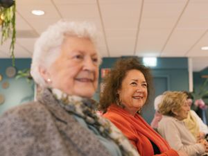 Een oudere dame met grijs jasje en net sjaaltje zit naast een vrouw met rode trui en gouden oorbellen. Ze kijken vrolijk voor zich uit.