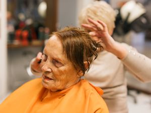 Dame met oranje kapmantel om en roodbruin nat haar wordt geknipt door dame met blond goedgekapt haar