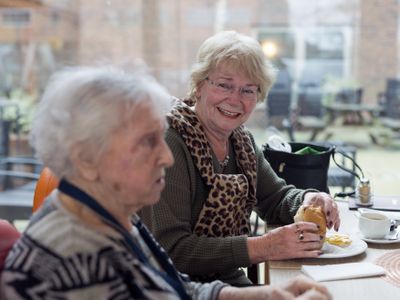 Twee dames met grijs haar zitten aan tafel met een kadetje in de hand, de ene dame kijkt vriendelijk naar de oudere dame