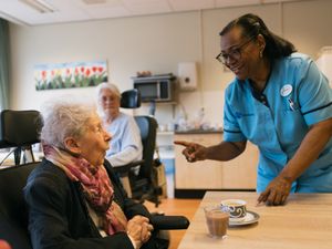 Een dame met kort grijs haar en kleurige sjaal zit in een rolstoel aan tafel met een kopje koffie. Een dame met zwart haar en bril in zorguniform kijkt haar lachend aan.