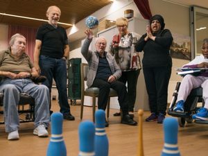 Een man op een stoel gooit een bal naar blauwe kegels op de grond. Om hem heen staan mensen mee te kijken en te klappen.