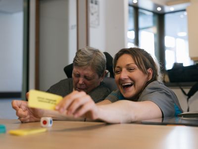 Jonge vrouw met bruin haar ondersteunt een zittende dame in rolstoel bij spelen van een spelletje aan tafel.